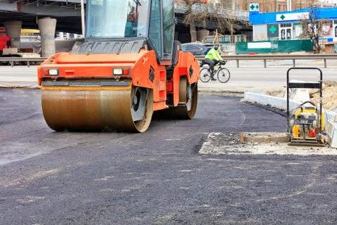 A heavy vibratory roller compacts hot asphalt on the roadway on a clear sunny Stock Photos
