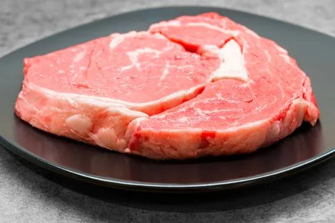High angle studio shot of a raw beef rib eye steak on a black design plate Stock Photos