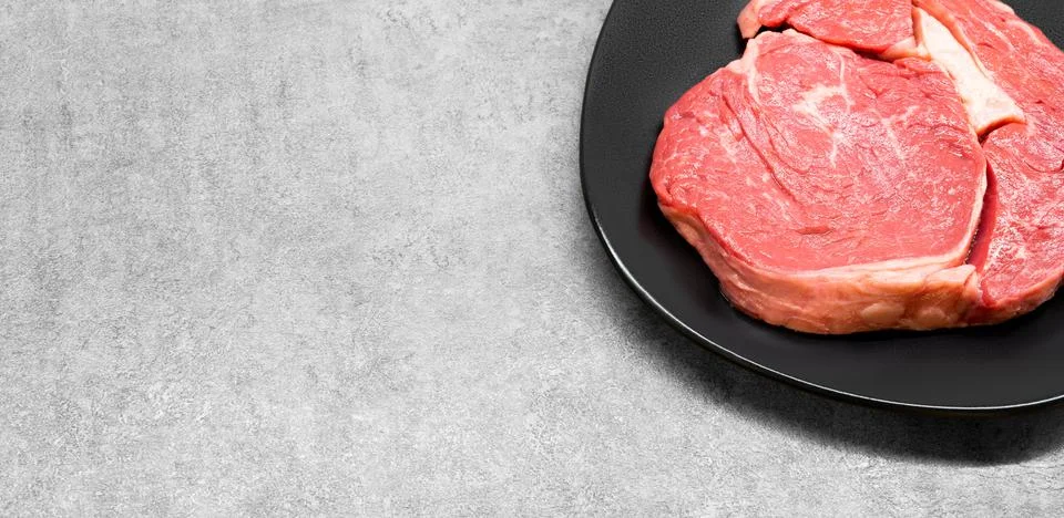 High angle studio shot of a raw beef rib eye steak on a black design plate Stock Photos