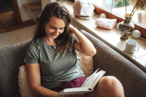 High angle view of lovely young woman with brown hair reading book on sofa at Stock Photos