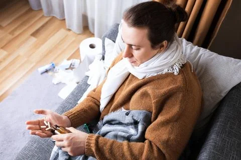 High angle view of sick man with scarf sitting on sofa pouring pills from b.. 스톡 사진
