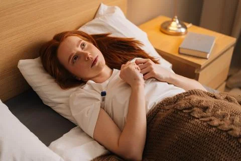 High-angle view of sick young woman with flu lying under blanket on bed holding Stock Photos