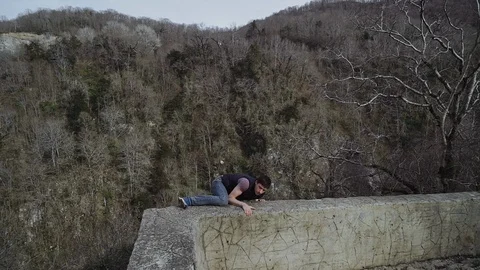 High in mountains a guy climbs onto a concrete fence, looks over and goes away 스톡 동영상 113672830