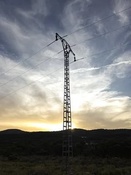 High voltage power line pylon stands against sunset sky over rural mountains Stock Photos