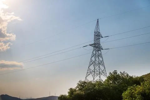 High voltage transmission lines tower over forested hillside under bright sun Stock Photos