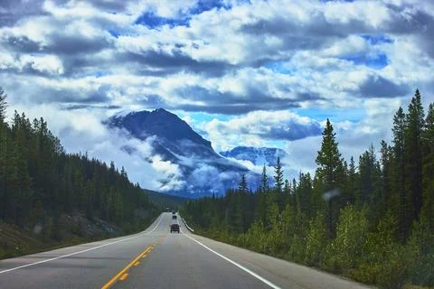 Highway Road Through Pine Forest With Dramatic Clouds And Snowy Mountain View 写真素材