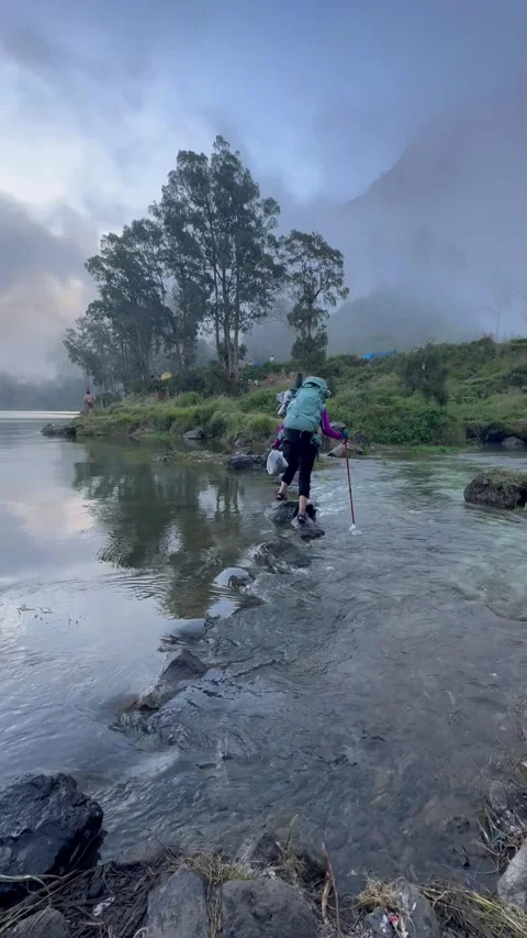 Hiker with backpack crossing a shallow stream on rocky path in misty mountain. Stock Footage 332258422