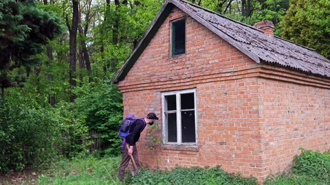 Hiker with backpack peering into the window of an old brick cabin Stock Footage 336970652
