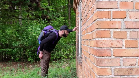 Hiker with backpack peering into the window of an old brick cabin Stock Footage 336970750