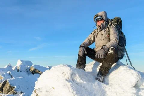 Hiker with backpack sitting and having rest on the top of a snow-covered rock Stock Photos