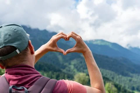 Hiker in cap with backpack forms heart shape with their hands while admiring Stock Photos