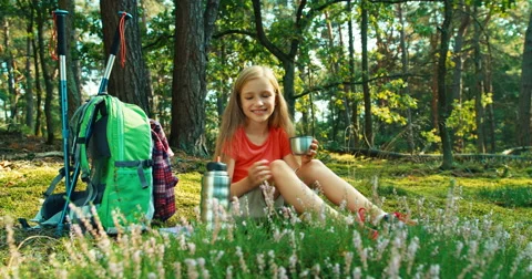 Hiker girl 8-9 years pours tea from a thermos sitting on the grass in forest Stock Footage