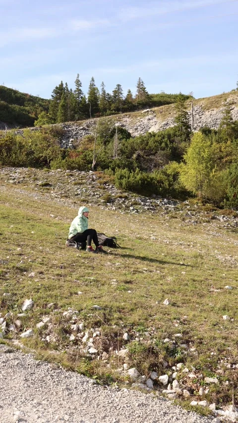 Hiker in green jacket sits on grassy rocky slope with backpack resting beside en Stock Footage 318219858
