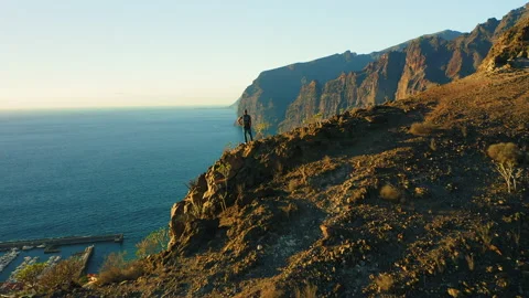 Hiker man with backpack on edge of sheer cliff overlooking sea waters. Mountain Video stock 230050469