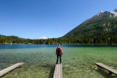 A hiker with an orange jacket and backpack stands at the end of a wooden pi.. Stock Photos