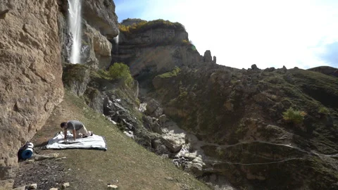 Hiker Setting Up a Tent at the Base of a Rock with Waterfall in the Background Video stock 318833752