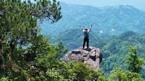 Hiker standing on mountain cliff edge with arms raised overlooking vast valley Video stock 342324589