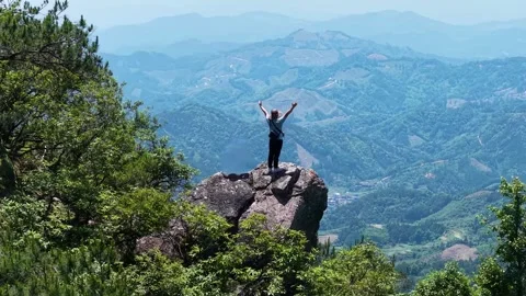 Hiker standing on mountain summit with arms raised overlooking valley landscape Stock Footage 342324579
