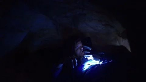 Hiker using smartphone laying on ground in closely hole in cave with flashlight Stock Footage 80062840