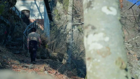 Hiker Walking Down Leaf-Covered Mountain Trail Towards Small Hermitage Stock Footage 316418490