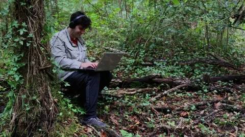Hiker works on a laptop in the forest while listening to music on headphones Stock Footage 289007760