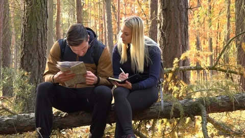 A hiking couple sits on a broken tree in a forest, the man reads off a map and Vídeos de archivo 109435715