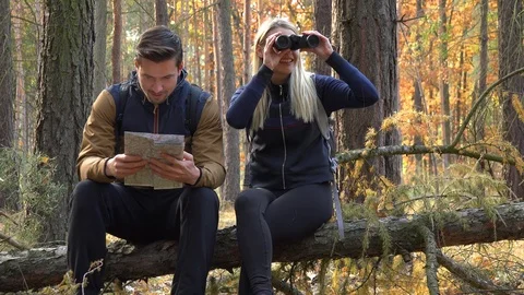 A hiking couple sits on a broken tree in a forest, the man reads off a map and Vídeos de archivo 109435725