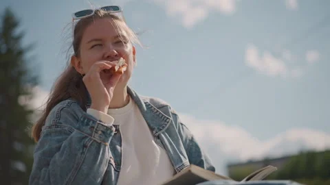 Hillside reading snack under sun, woman in denim jacket bites pastry with open Stock Footage 346544984