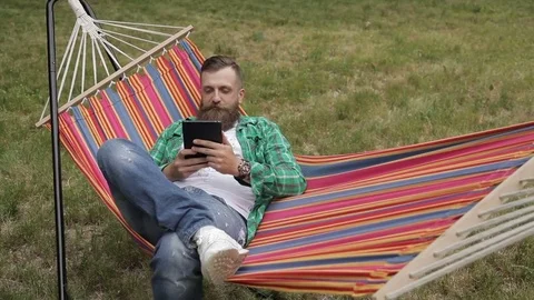 Hipster with a beard resting on a hammock with a tablet in hands Stock Footage 77167488