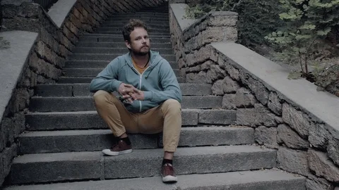 Hipster guy with a beard sits on the stone steps on the stone stairs and smiles Stock Footage 115480536