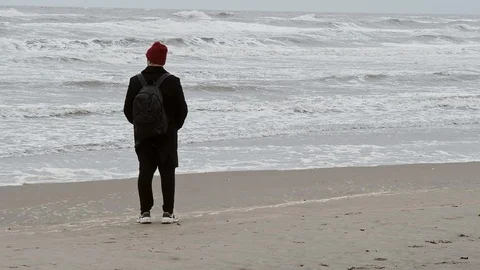Hipster man in red hat standing alon and walking across the beach in autumn. 스톡 동영상 119638685