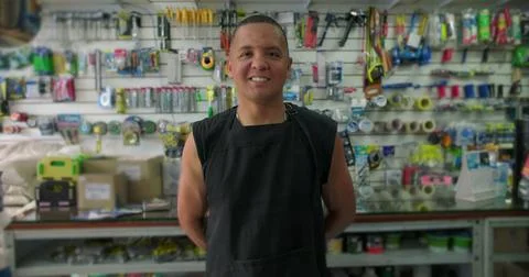 Hispanic hardware store worker smiles gently in shop aisle expressing pride.. Stock Photos