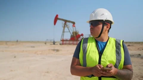 Hispanic male technician checking mobile data on phone by pumpjack, wearing whi Stock Footage 326802391