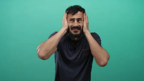Hispanic man with beard in distress holding head against vibrant green back.. 스톡 동영상 314254737