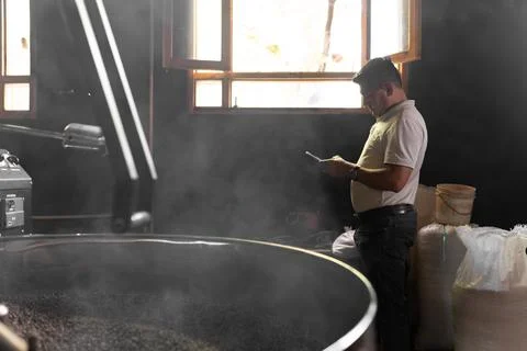 An Hispanic man is checking his phone next to a coffee roaster machine Stock Photos