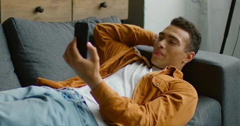 Hispanic man is lying on sofa, having a video chat via smartphone 스톡 동영상 114643335