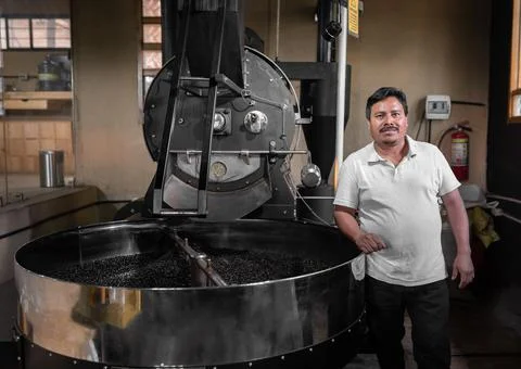 A Hispanic man is posing next to his big coffee roaster machine Stock Photos