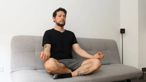 Hispanic man sitting at the left side of a couch in the lotus pose meditating Видео 340583423