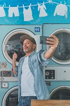 Hispanic man taking selfie while doing laundry at laundromat Stock Photos