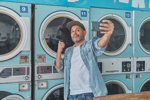 Hispanic man taking selfie while doing laundry at laundromat Stock Photos