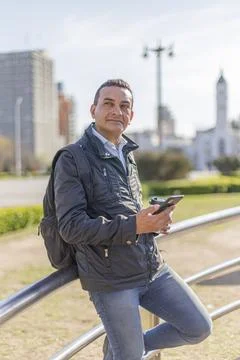 Hispanic man using his mobile phone leaning on a railing in a public park. Photos