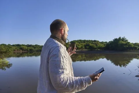 Hispanic man using his smartphone while he drinks mate outdoors, in nature. Stock Photos