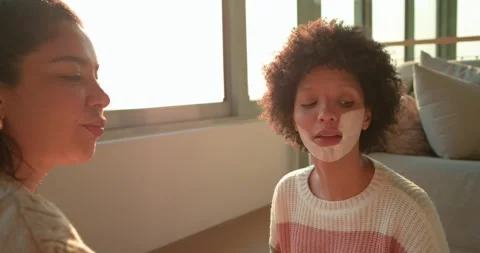 Hispanic mom brushing mask onto daughter's cheeks for pampering in living room, Stock Footage 335808583