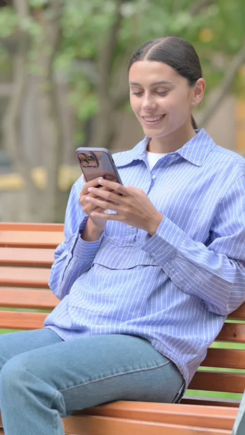 Hispanic Woman using Smartphone while Sitting Outdoor, vertical Stockbeeldmateriaal 321121567