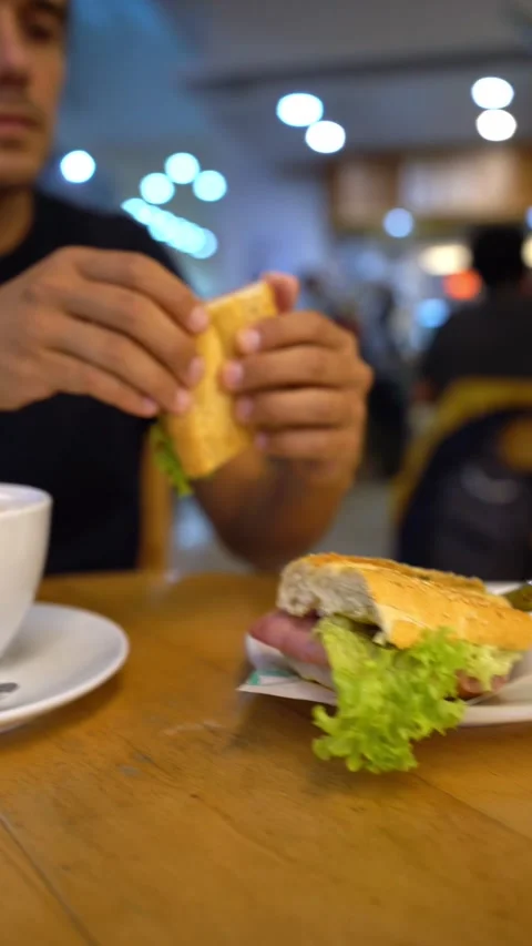 Hispanic young man eats a ham and cheese sandwich in a coffee shop Video stock 163367309