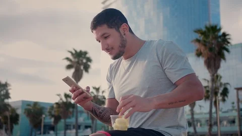 Hispanic young man using new application on mobile phone and eating fruits at Stock Footage 108443719