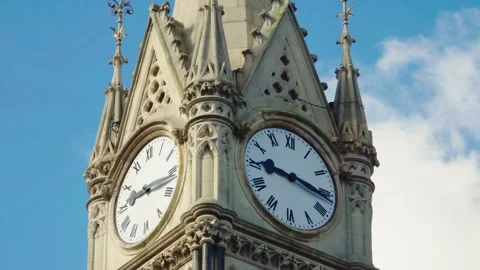 Historic gothic clock tower with roman numeral faces against blue sky 스톡 동영상 335144203