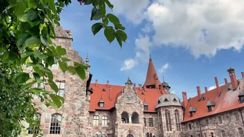 Historic Stone Castle Building With Red Tile Roof and Turrets Framed by Green Stock Footage 346615668
