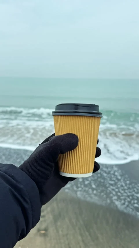 Holding a coffee cup by the shore with waves in the background on a chilly day Stock Footage 341601683