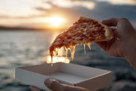 Holding a pizza slice above an empty box at sunset by the water Fotos Stock